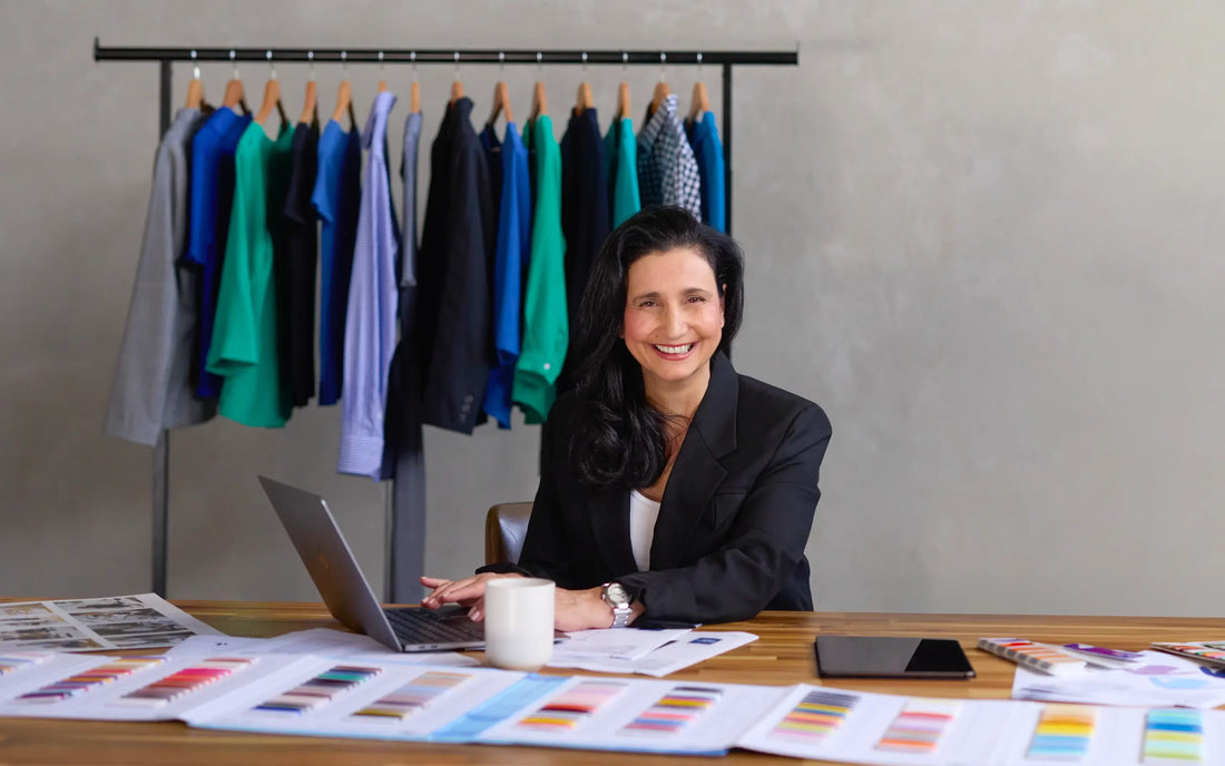 Maria Grossi, CEO and Founder of Designs To You, sitting at a desk with fabric swatches and documents, smiling at the camera. In the background, various colourful garments are hanging on a rack.