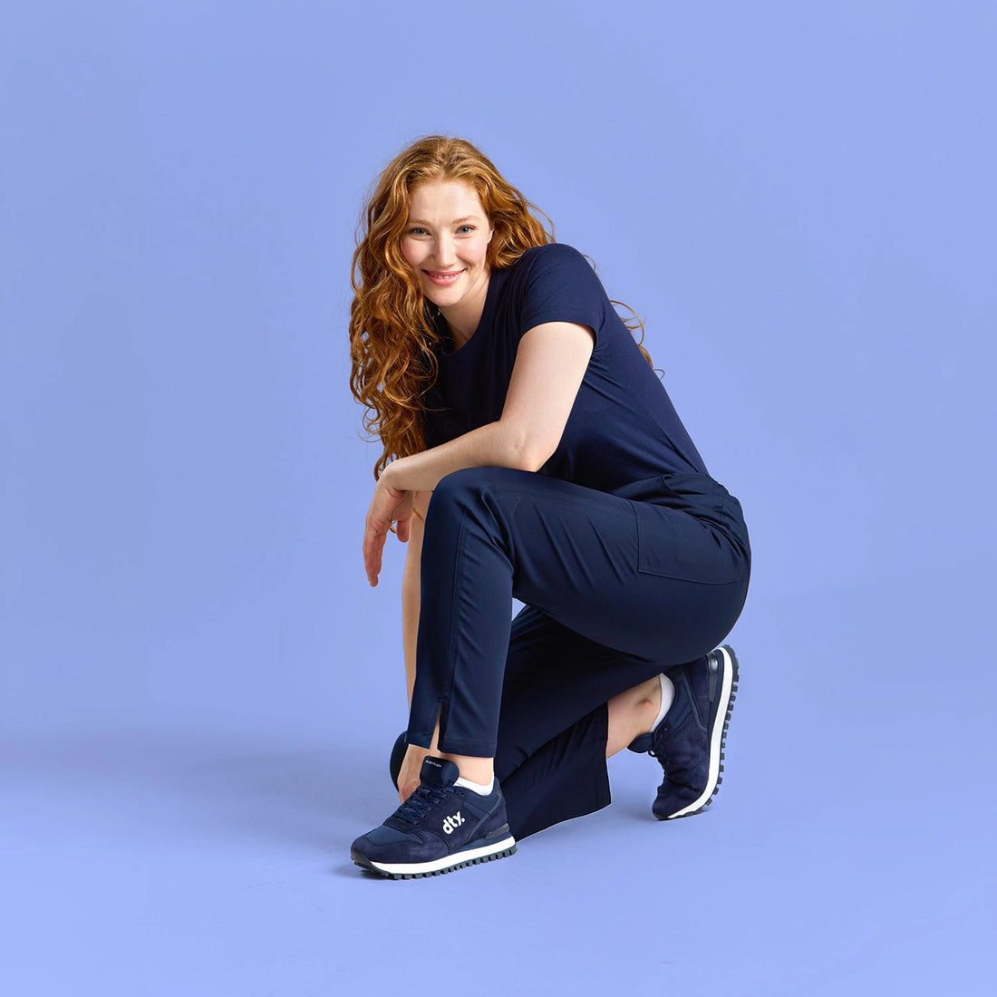 Woman wearing navy medical scrubs and DTY-branded sneakers, crouching confidently against a matching blue studio background.