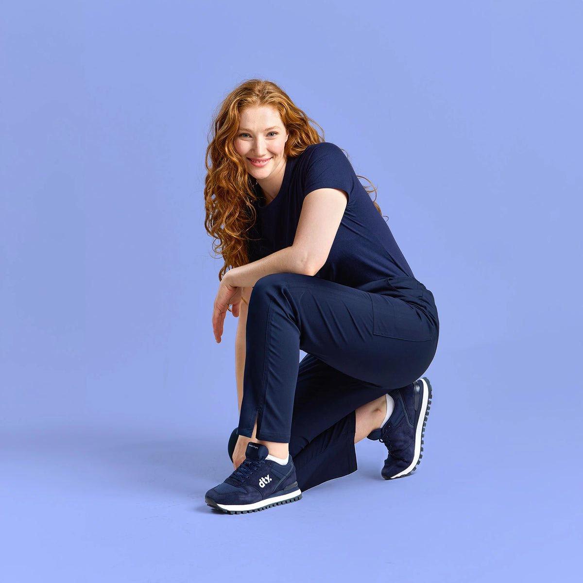 Woman wearing navy medical scrubs and DTY-branded sneakers, crouching confidently against a matching blue studio background.
