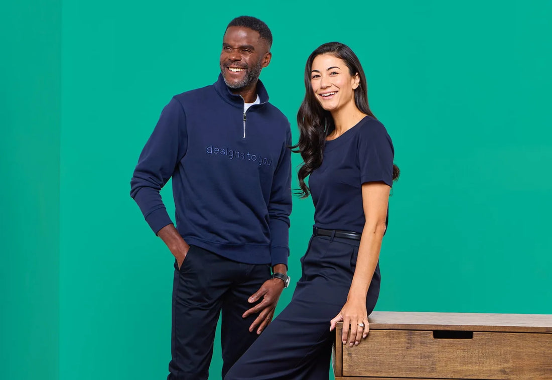 Man and woman in navy blue corporate uniforms pose against a green background. The man wears a quarter-zip pullover with 'designs to you' embroidery, while the woman wears a short-sleeved blouse with tailored trousers.