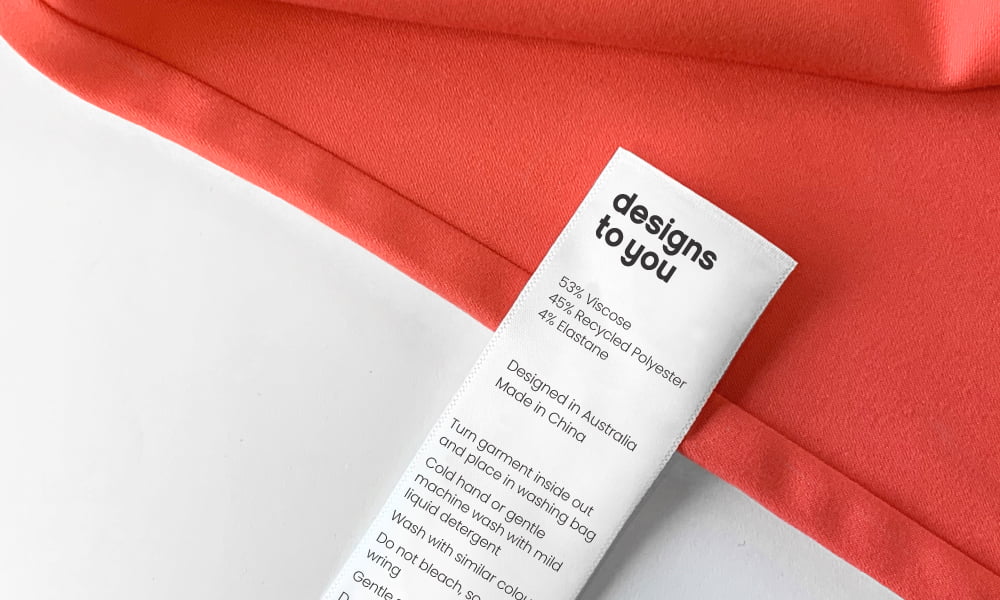 The banner for the article 'Your Care Label is Trying to Tell You Something' displays a white care label for a clothing item manufactured for Designs To You. The background features a watermelon coloured fabric sitting atop a plain white background.