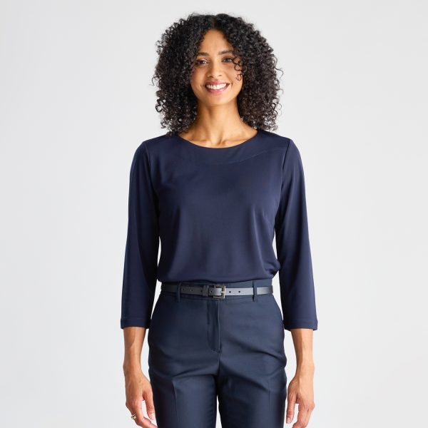 a Smiling Woman with Curly Hair Models a Navy Soft Knit 3/4 Sleeve Boat Neck Top and Trousers, Paired with a Black Belt and Standing Against a White Background.