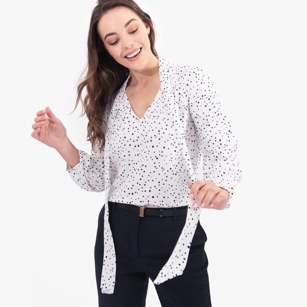 a Woman Smiling, Wearing a White V-neck Tie-front Blouse with Navy Polka Dots, Playfully Holding the Tie Fronts.