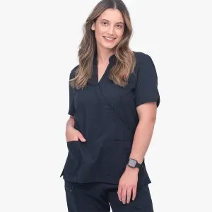 Woman Wearing a Twilight Navy Scrub Top with Crossover Front and Short Sleeves, Standing with Hands in Pockets and Smiling at the Camera.