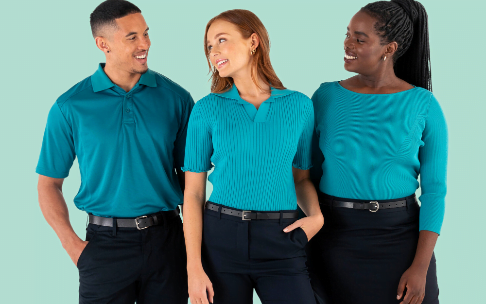 The banner for the article 'Sustainable Uniforms: The Essential Guide for Procurement Managers' depicts two female and one male model posing together and smiling for the camera in front of a aqua coloured background. They're wearing modern and stylish teal coloured uniforms manufactured by Designs To You. The colour palette of the photo reflects the blog topic - sustainable uniforms.