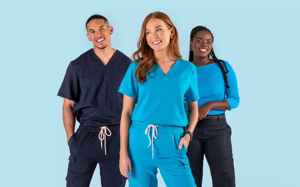 The banner for the article 'Making Your Uniform Program More Sustainable' depicts three young, professional looking models wearing stylish, high-quality blue and navy healthcare uniforms manufactured by Designs To You. They're smiling confidently at the camera and standing in front of a a light blue background.
