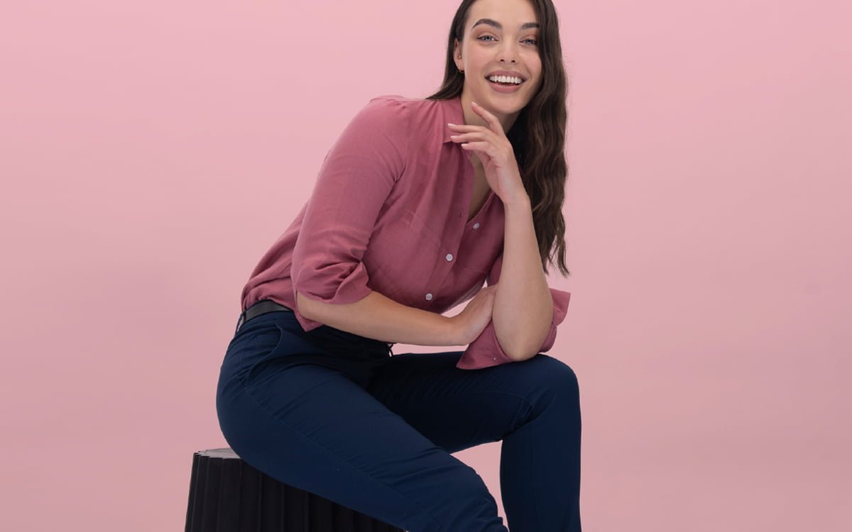 The banner for the article 'The Real ROI of Work Uniforms' depicts a professional-looking female wearing a pink linen, button-up blouse and navy slacks manufactured by Designs To You. She is sitting atop a black chair and smiling radiantly at the camera in front of a pink background.