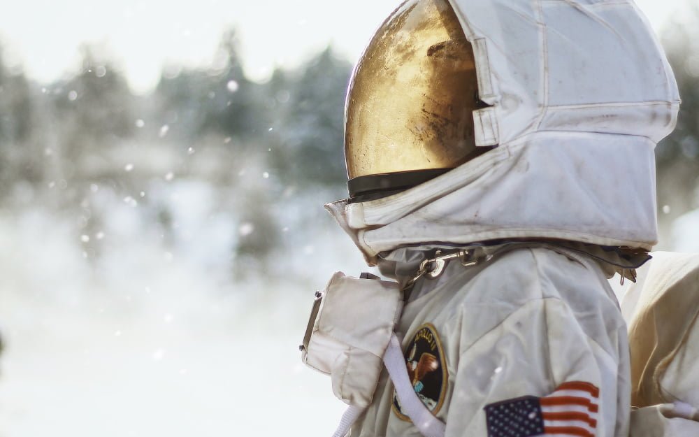 The banner for the article 'What Do Astronauts and Uniforms Have in Common?' features a United States astronaut wearing their space suit, faced parallel to the camera. The blurry background looks to be a snowy, outdoor winter landscape.
