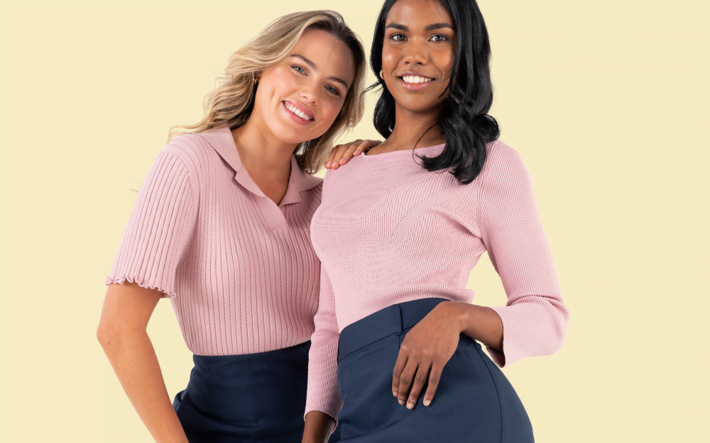 The banner for the article 'Why Smaller Uniform Suppliers Reign Supreme' depicts two female models posing together and smiling for the camera in front of a pale yellow background. They're wearing modern, professional and stylish light pink tops manufactured by Designs To You.