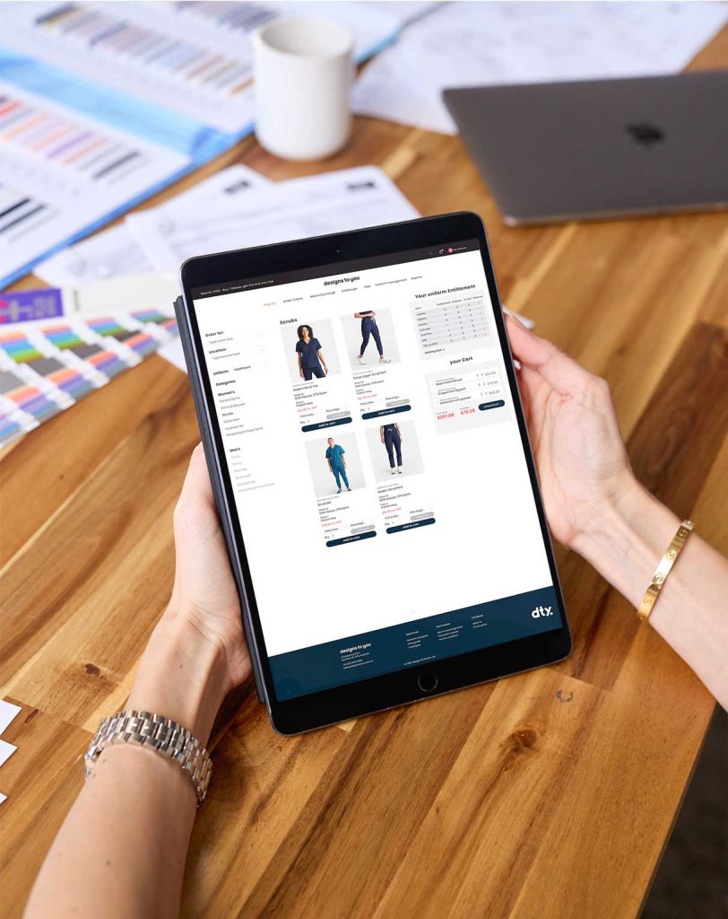 Online Uniform Solutions Management System: A photo of a black iPad being held in front of somebody with Designs To You's Online Uniform Management System visible on the screen. Papers, a coffee mug and a laptop can be seen sitting on a wooden table in the background. 