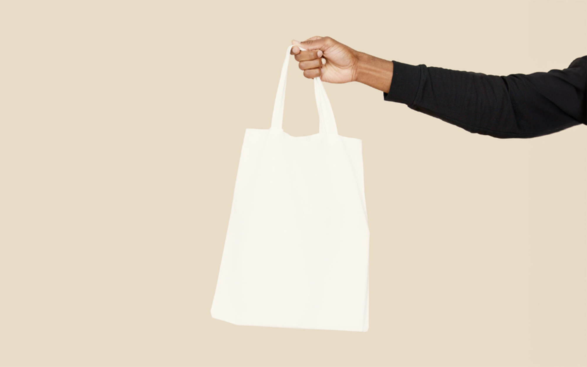 A person's arm is shown extending from the right side of the image, holding a white tote bag against a neutral background. The tote bag is a product made from recycled uniforms.