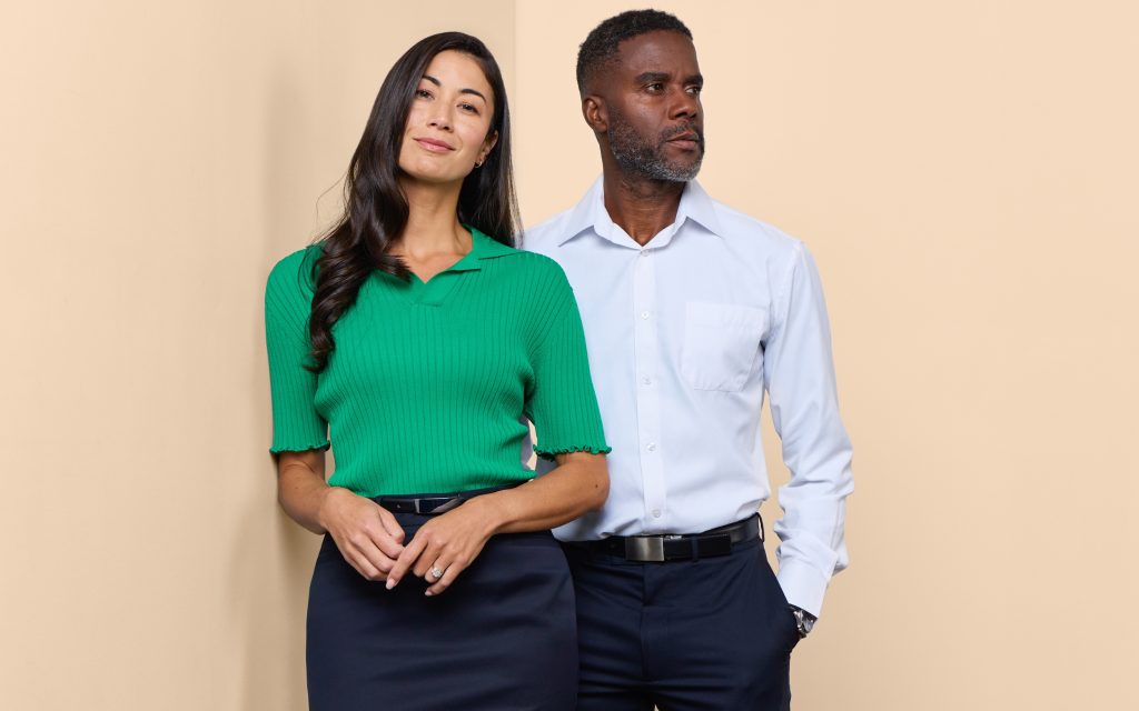 a Man and Woman Standing Side by Side Against a Beige Background, the Woman to the Left Wearing a Green Ribbed Polo Shirt and Navy Skirt, the Man to the Right in a Light Blue Button-up Shirt and Dark Trousers.