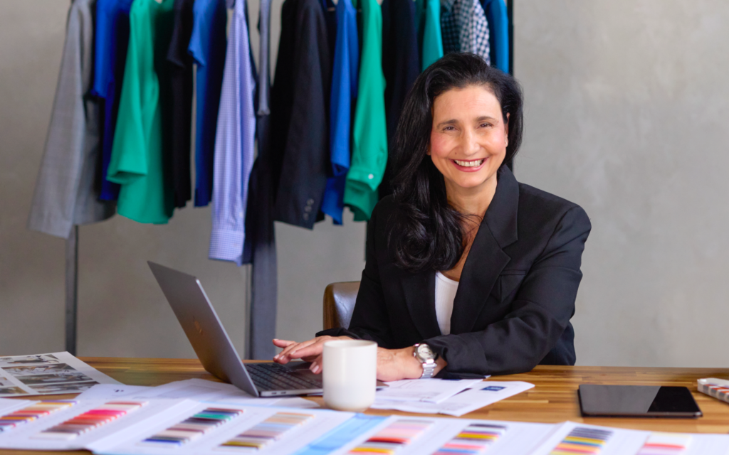 Maria Grossi, Ceo and Founder of Designs to You, Sitting at a Desk with Fabric Swatches and Documents, Smiling at the Camera. in the Background, Various Colourful Garments Are Hanging on a Rack.