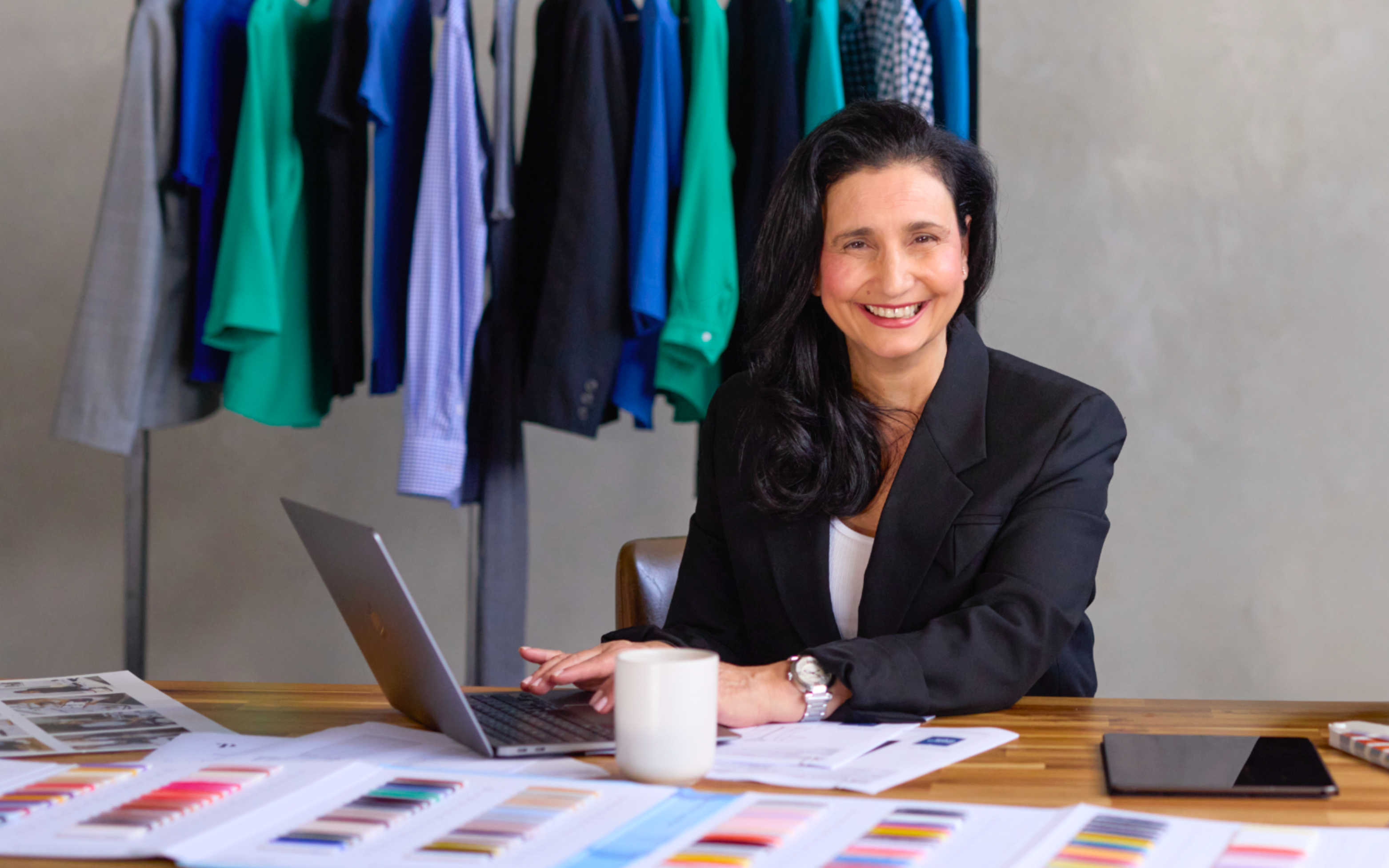 Maria Grossi, CEO and Founder of Designs To You, sitting at a desk with fabric swatches and documents, smiling at the camera. In the background, various colourful garments are hanging on a rack.