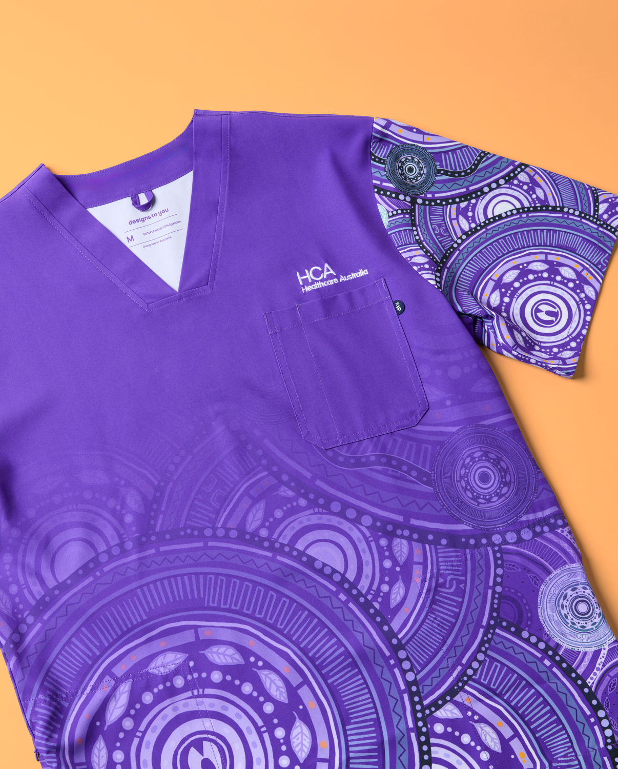 a Vibrant Purple Scrub Top is Displayed Against an Orange Background. the Scrub Top Features a V-neck Design with Intricate First Nations-inspired Artwork Sublimated on the Sleeves and Lower Half of the Garment. the Hca Logo is Embroidered on the Chest Pocket. the Detailed Patterns and Rich Purple Colour Make the Top Visually Striking and Culturally Significant.