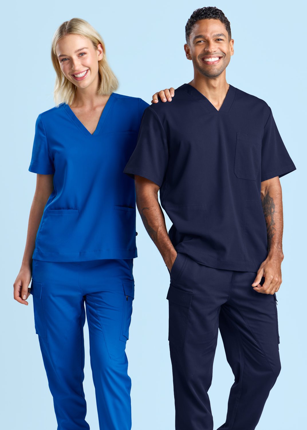 Two Healthcare Professionals Standing Side by Side, One Wearing Vibrant Blue Scrubs with a V-neck Top and Matching Cargo Pants, and the Other in Deep Navy Scrubs with a Chest Pocket and Cargo Pants, Both Smiling Against a Light Blue Background.