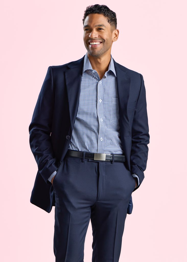 a Smiling Man Dressed in a Corporate Navy Suit with a Gingham Check Shirt, Looking Confident and Approachable Against a Soft Pink Background.