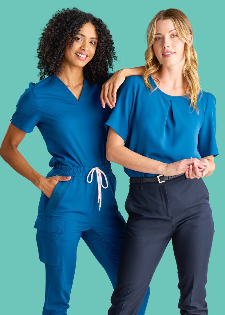 Two Healthcare Professionals in Coordinating Blue Scrubs, One with Curly Hair and the Other with Blonde Hair, Standing Confidently Together Against a Light Green Background.