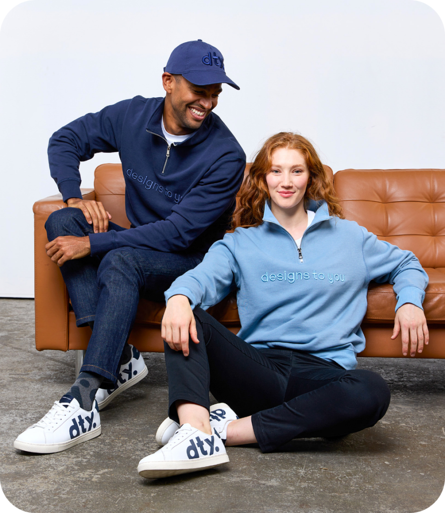 a Man and a Woman Sitting on the Floor and Couch, Both Wearing Custom Blue and Navy "designs to You" Branded Sweatshirts and Matching Sneakers, Smiling and Looking Relaxed.