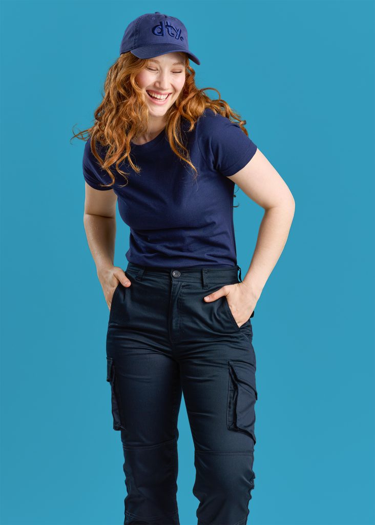 a Cheerful Woman in Navy Blue Workwear, Including a T-shirt and Cargo Pants, Topped with a Matching Cap Embroidered with "dty." She is Laughing and Looking to the Side Against a Light Blue Background.