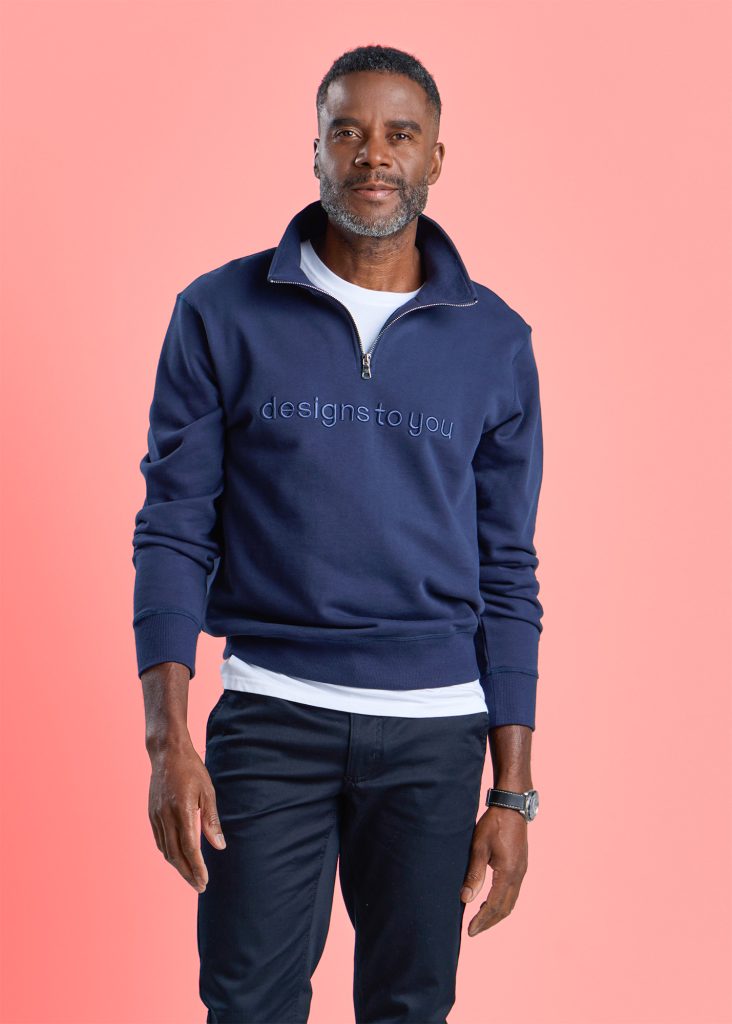 a Man in a Navy Blue Half-zip Pullover Embroidered with "designs to You," Paired with Dark Trousers, Standing Confidently Against a Soft Pink Background. His Expression is Calm and Composed.