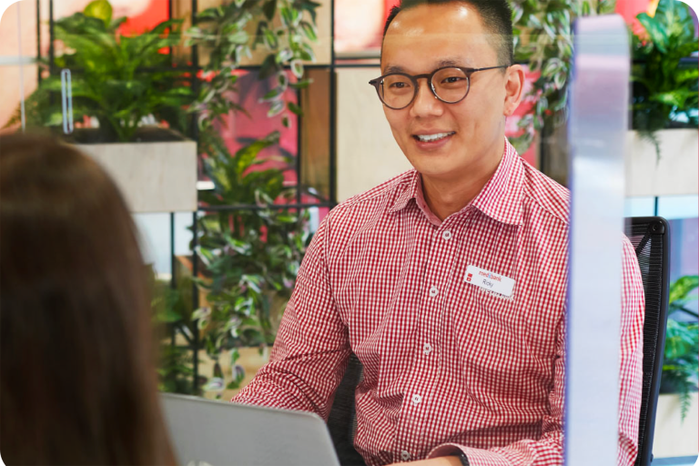 A Medibank employee, Ricky, sits at a desk wearing a red and white checkered shirt, engaging in conversation with a customer across the table. The background features lush green plants, creating a welcoming environment.