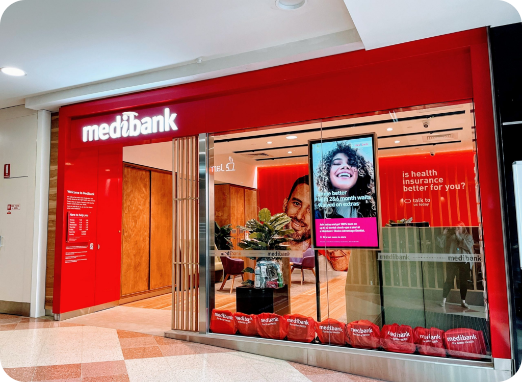 the Exterior of a Medibank Retail Store, Featuring the Prominent Medibank Logo in White Against a Red Background. the Store Has Large Glass Windows Displaying Vibrant Promotional Materials and Plants Inside. a Digital Signboard Shows a Smiling Customer, Reinforcing Medibank's Commitment to Customer Satisfaction and Health Insurance Services.