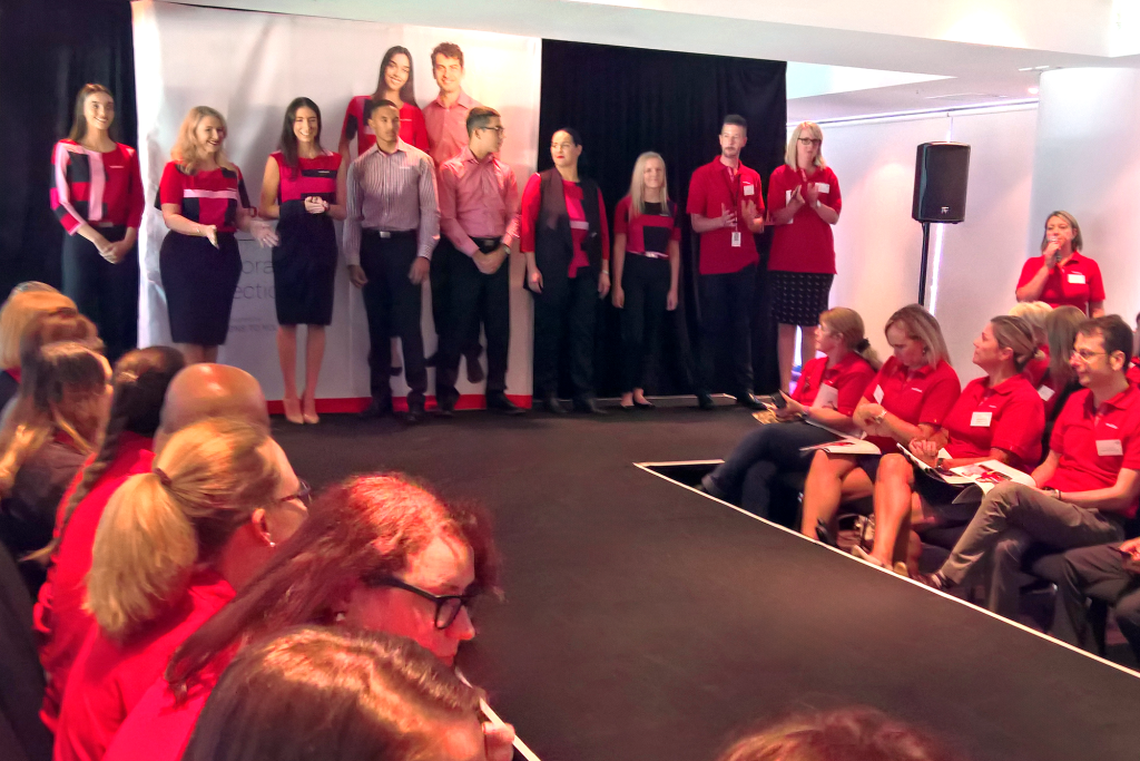 the Image Shows a Group of People Standing on a Stage During a National Uniform Roadshow for Medibank's New Uniform Collection in 2017. the Individuals on Stage Are Wearing the New Uniforms, and the Audience, Seated in Front, is Attentively Watching the Presentation.