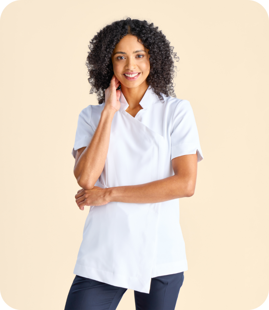 a Woman Wearing a White Short-sleeve Wrap-style Tunic, Smiling and Posing with One Hand Near Her Face. She Has Curly Hair and is Standing Against a Plain Light Background.