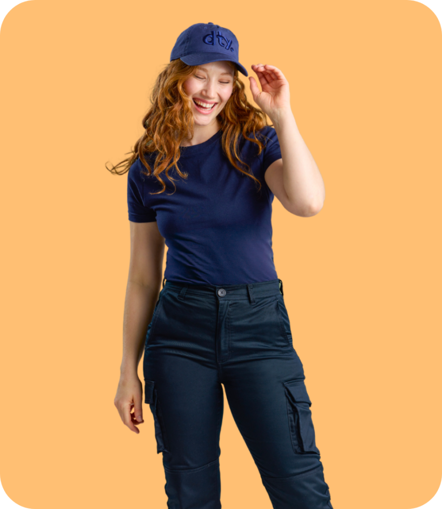 a Person with Wavy Red Hair Wearing a Navy Blue T-shirt, Matching Cargo Pants, and a Cap with the "dty" Logo, Part of Designs to You's Workwear Uniforms Collection. the Background is a Solid Light Orange, Creating a Vibrant Contrast with the Uniform. the Person is Smiling and Adjusting the Cap, Showcasing the Comfortable and Stylish Design of the Workwear.