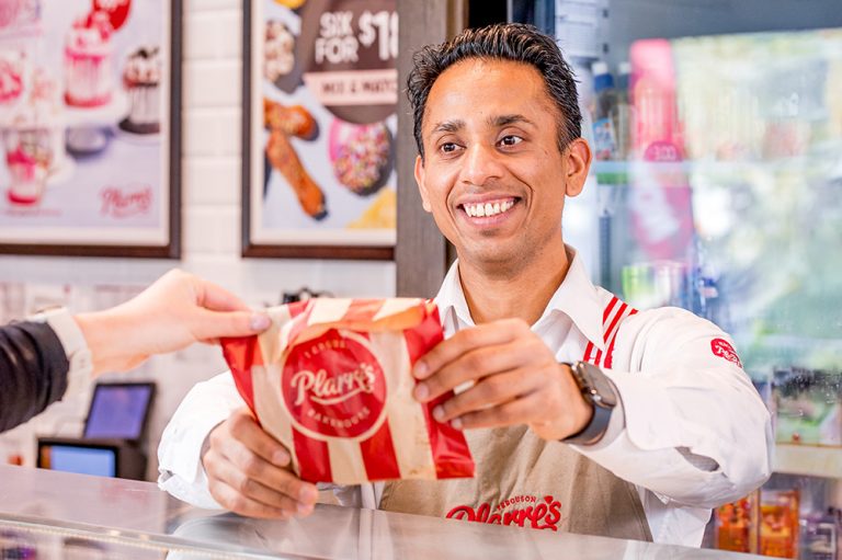 A smiling Ferguson Plarre team member hands a customer a red and white striped package over the counter, wearing the brand's signature apron with red straps.