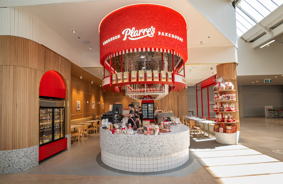 Interior view of Ferguson Plarre Bakehouse featuring a prominent red cylindrical counter with the bakery's logo, surrounded by wooden panel walls, display shelves filled with bakery goods, and a seating area.