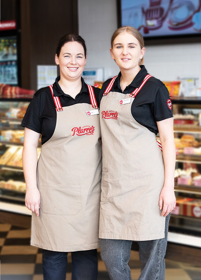 Two Smiling Team Members at Ferguson Plarre's Bakehouse Wearing Beige Aprons with Red and White Striped Straps, Standing in Front of a Bakery Display Case.