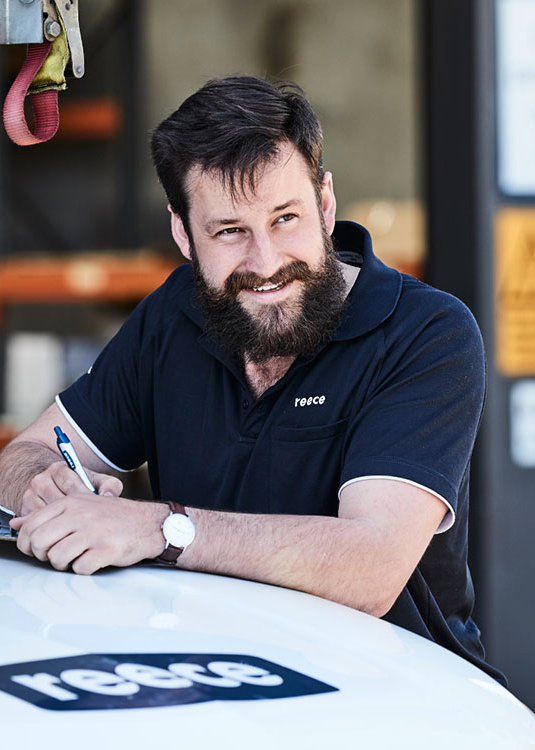 a Man with a Full Beard, Smiling Warmly, Leaning on a Vehicle Marked with the Reece Logo. He's Wearing a Navy Reece Polo Shirt and is Holding a Pen, Possibly Taking Notes or Filling out a Form.