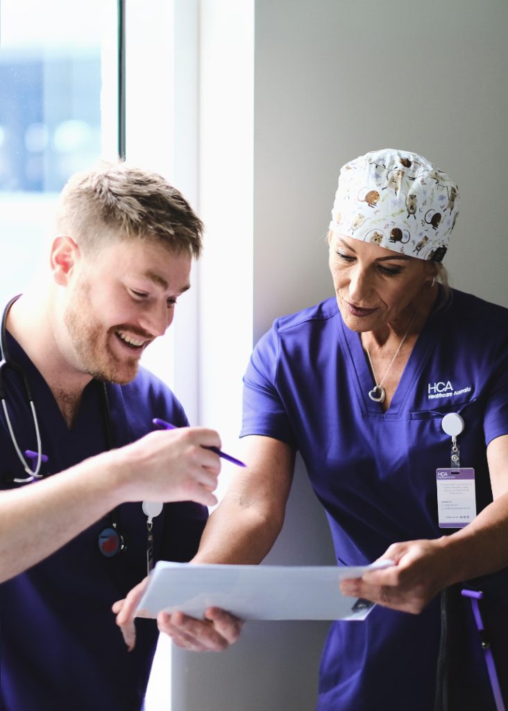 a Male and Female Healthcare Professional, Both in Scrubs, Engaging in a Discussion over a Clipboard. the Woman is Wearing a Scrub Cap with a Playful Print and a Navy Blue Scrub Marked with the "hca Healthcare Australia" Logo, While the Man in a Darker Scrub Smiles As He Writes.