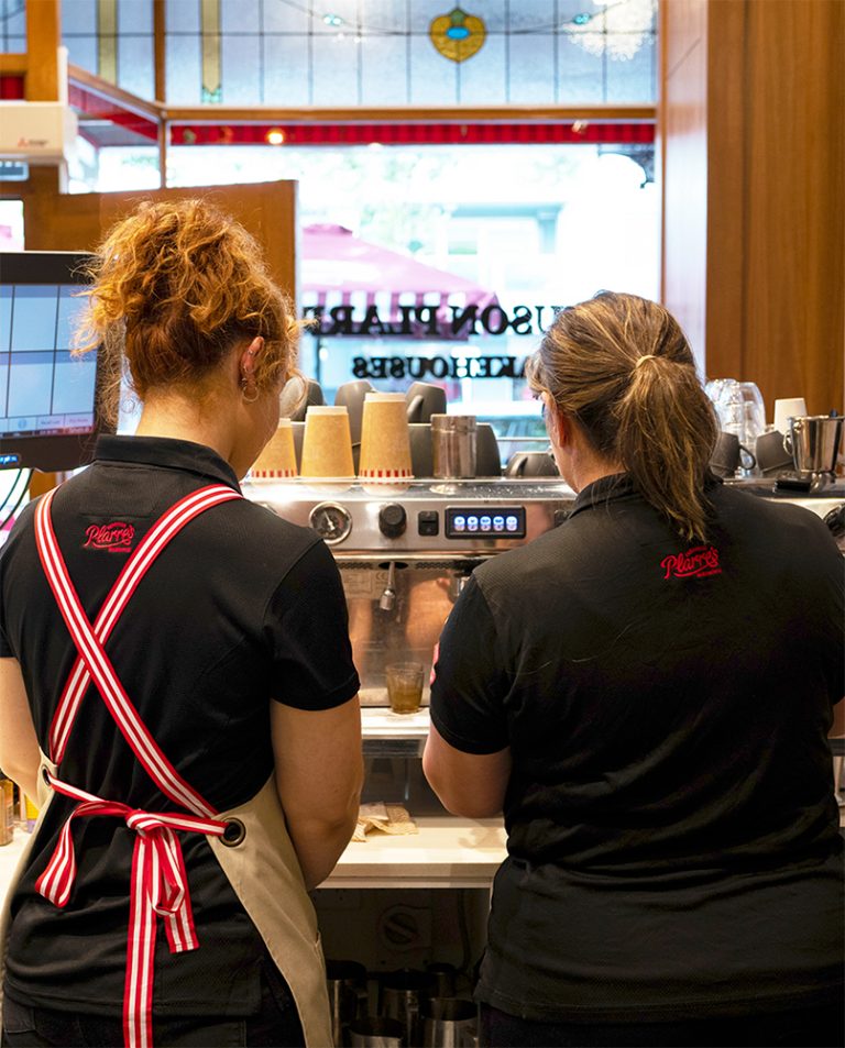 Two Ferguson Plarre Team Members, Seen from Behind, Work at a Coffee Machine. One Wears a Beige Apron with Red Striped Straps, While the Other is in a Black Polo with the Brand’s Logo.