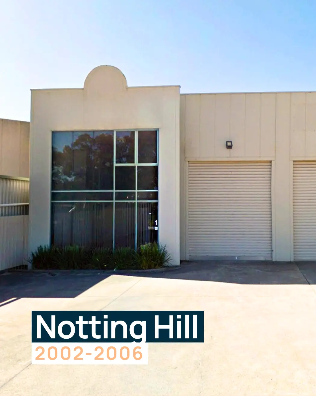 a Commercial Building with Large Glass Windows on the Left and Two Roller Doors on the Right. the Building Features a Minimalist Cream Exterior with a Distinct Arch Design at the Top. This Location Housed Designs to You During the Early 2000s.