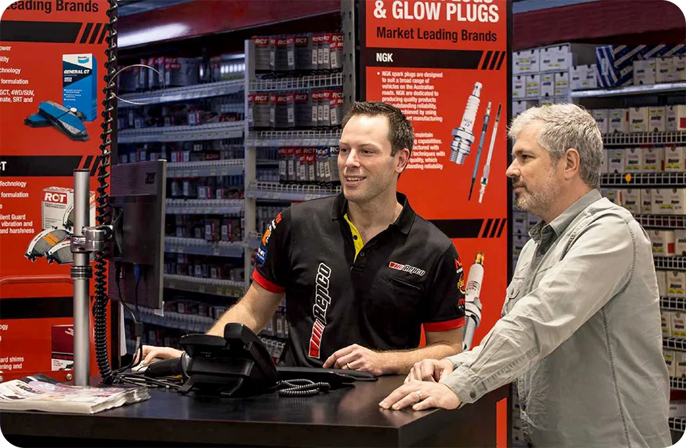 A male employee in a Repco uniform assisting a male customer at a service counter inside an auto parts store.