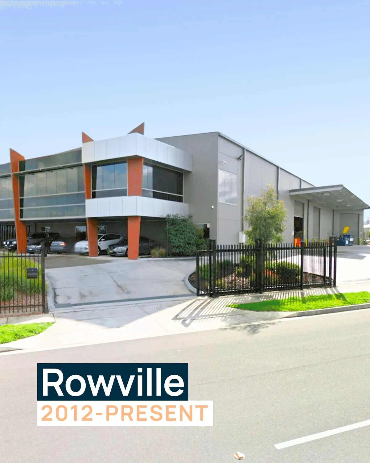a Large, Modern Industrial Building with an Orange and Grey Exterior. It Features Glass Windows on the Second Floor and a Secured Parking Lot at the Front. This Location Currently Serves As the Headquarters for Designs to You.