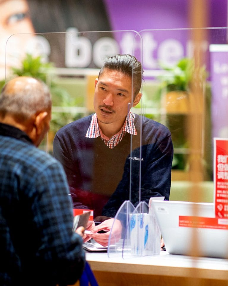a Medibank Customer Service Representative Wearing a Branded Uniform Sweater and Custom Collared Work Shirt, Assisting a Customer at a Service Desk in a Retail Setting.