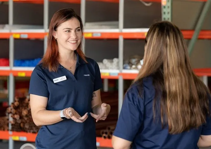 A Reece Group employee wearing a navy blue polo shirt and a name tag, smiling and engaging in a conversation with a colleague. They are standing in a warehouse or storage area with shelves stocked with materials in the background.