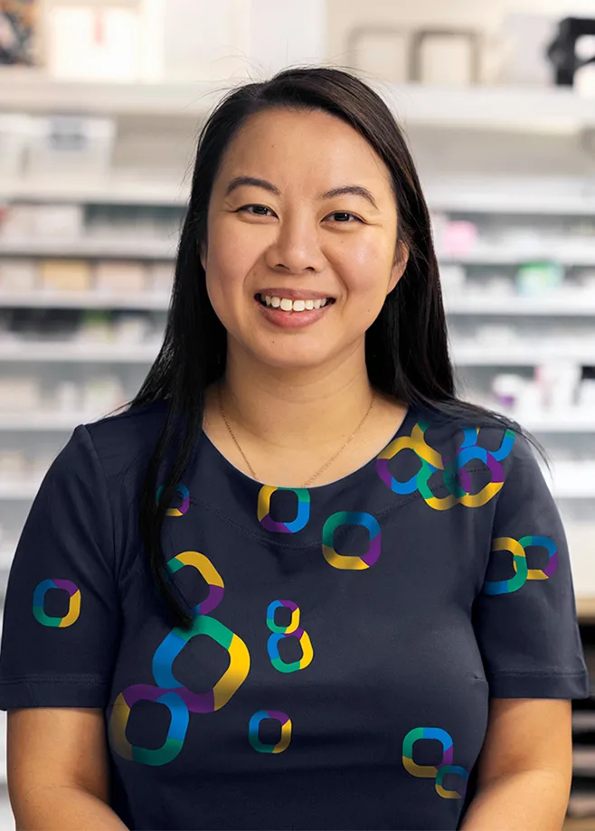 Smiling woman wearing a navy scrub top with colourful geometric patterns, seated in a modern pharmacy or medical workspace with shelves and office equipment in the background.