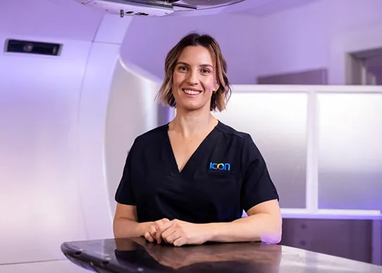 Smiling Female Healthcare Professional Wearing a Black Icon Group Scrub Top, Standing in a Medical Treatment Room with Equipment in the Background.