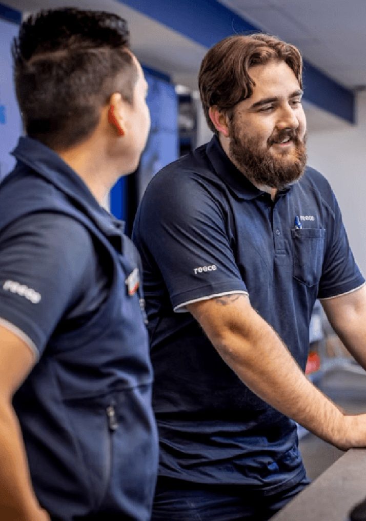 Two male Reece Group employees wearing navy blue uniforms. They are standing at a counter, engaged in conversation, with one facing the camera and the other turned slightly to the side. Both uniforms feature the Reece logo on the sleeves.
